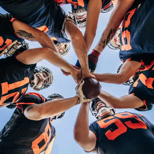 Low-angle view of American football players in uniform gathering in a team huddle, joining hands around a football before competition.