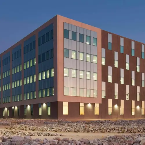 Exterior view of a modern corporate office building featuring large glass windows, architectural detailing, and illuminated workspaces at dusk.