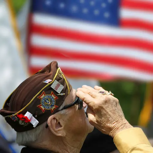 Elderly military veteran wearing a VFW cap raises a salute while facing an American flag.