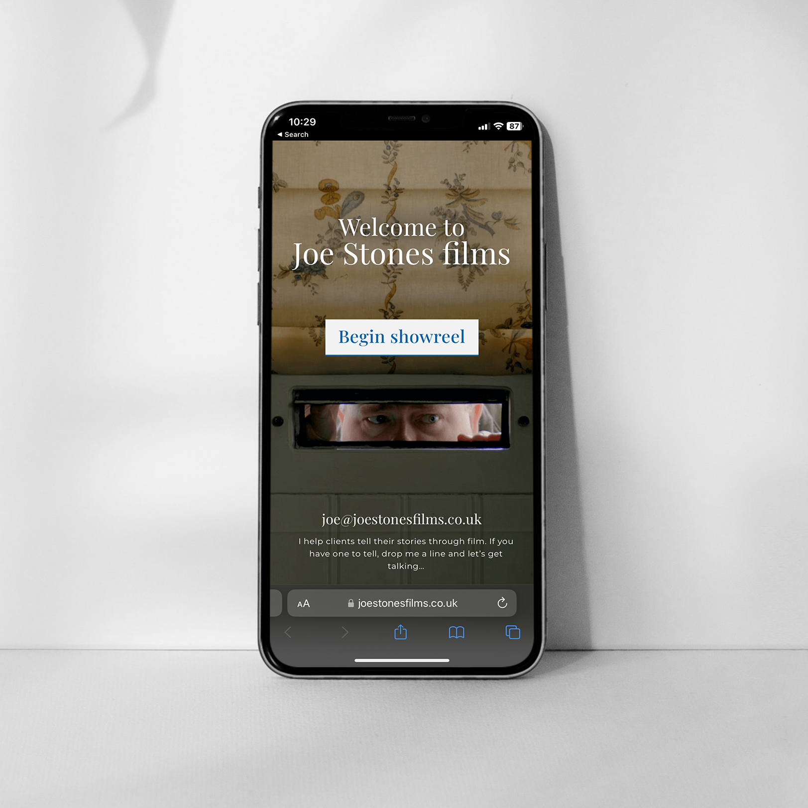 A mockup of a website design for a freelance filmmaker. A Apple iPhone standing upright on a white table, showing off the landing page of a beautiful light themed mobile website. The iPhone is leaning against a white wall.