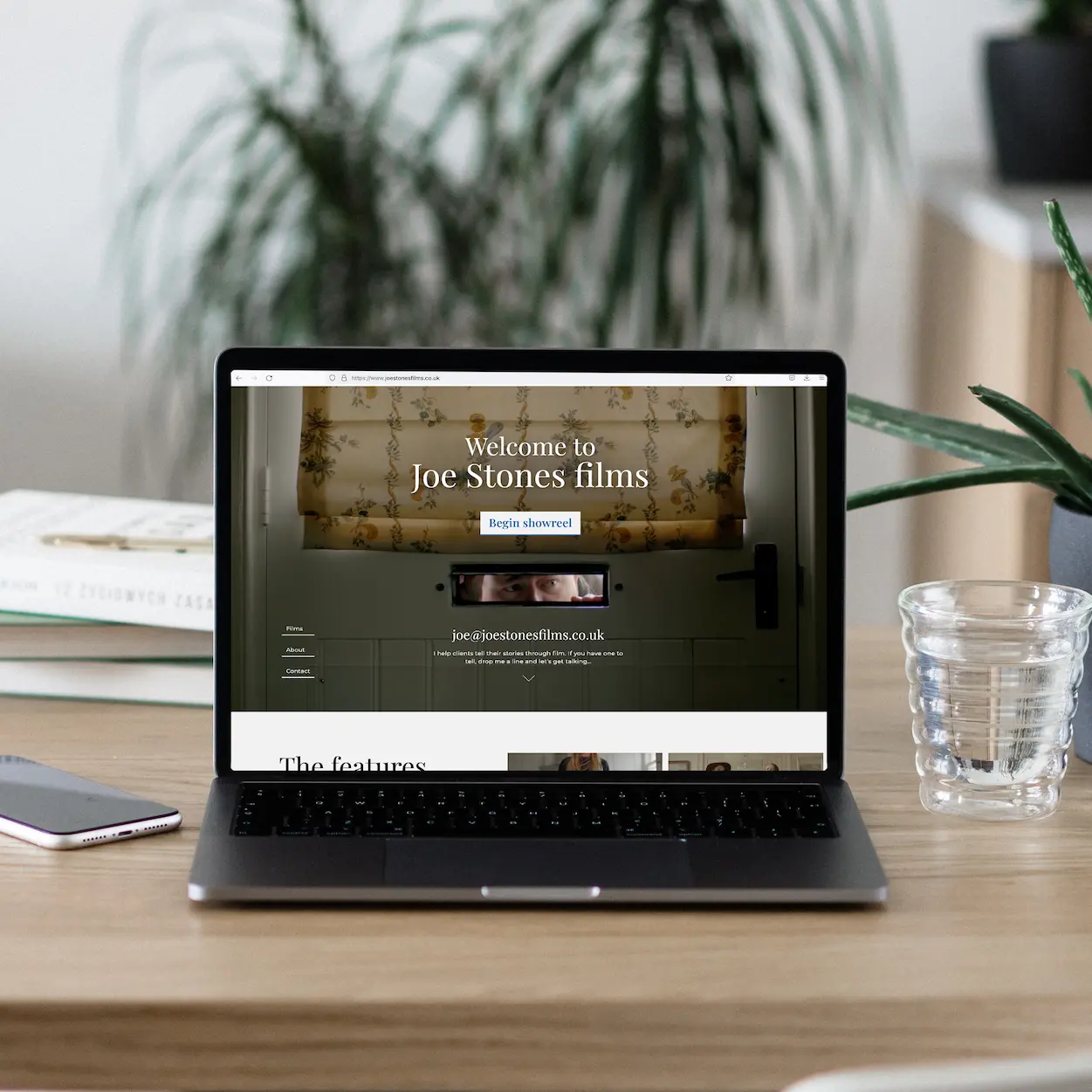 A mockup of a website design for a freelance filmmaker. A Macbook computer sitting on a wooden table, showing off the landing page of a beautiful light themed website. In the background there are deep green floor standing spikey plant.