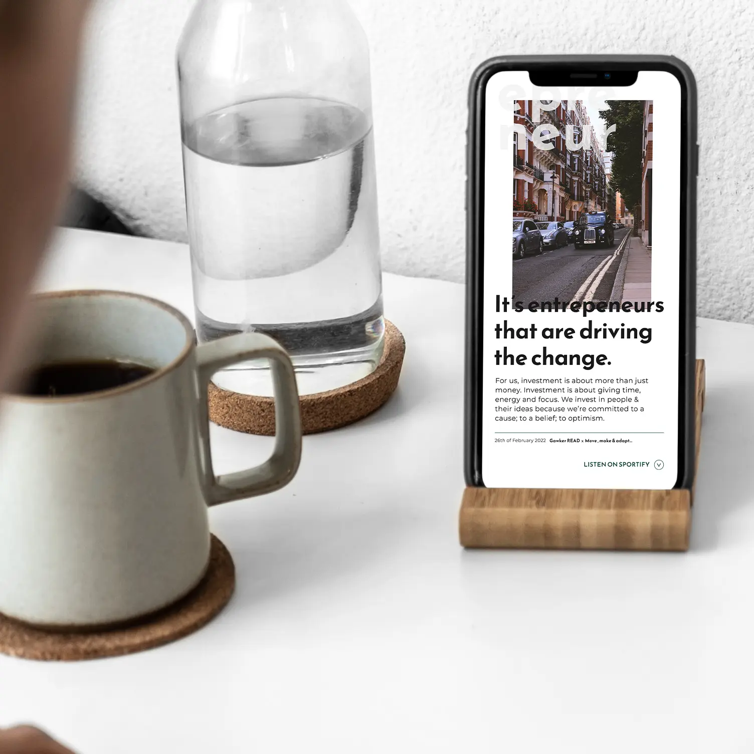 A mockup of a website design for a venture capital company. A Apple iPhone standing upright on a white table, showing off the about section of a beautiful light themed mobile website. The iPhone is surrounded by a bottle of water and a black coffee in a light grey mug.