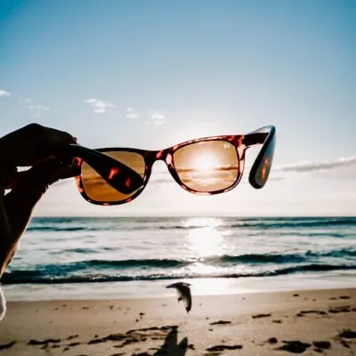 Hand holding sunglasses with sunset reflecting on lens over beach waves.