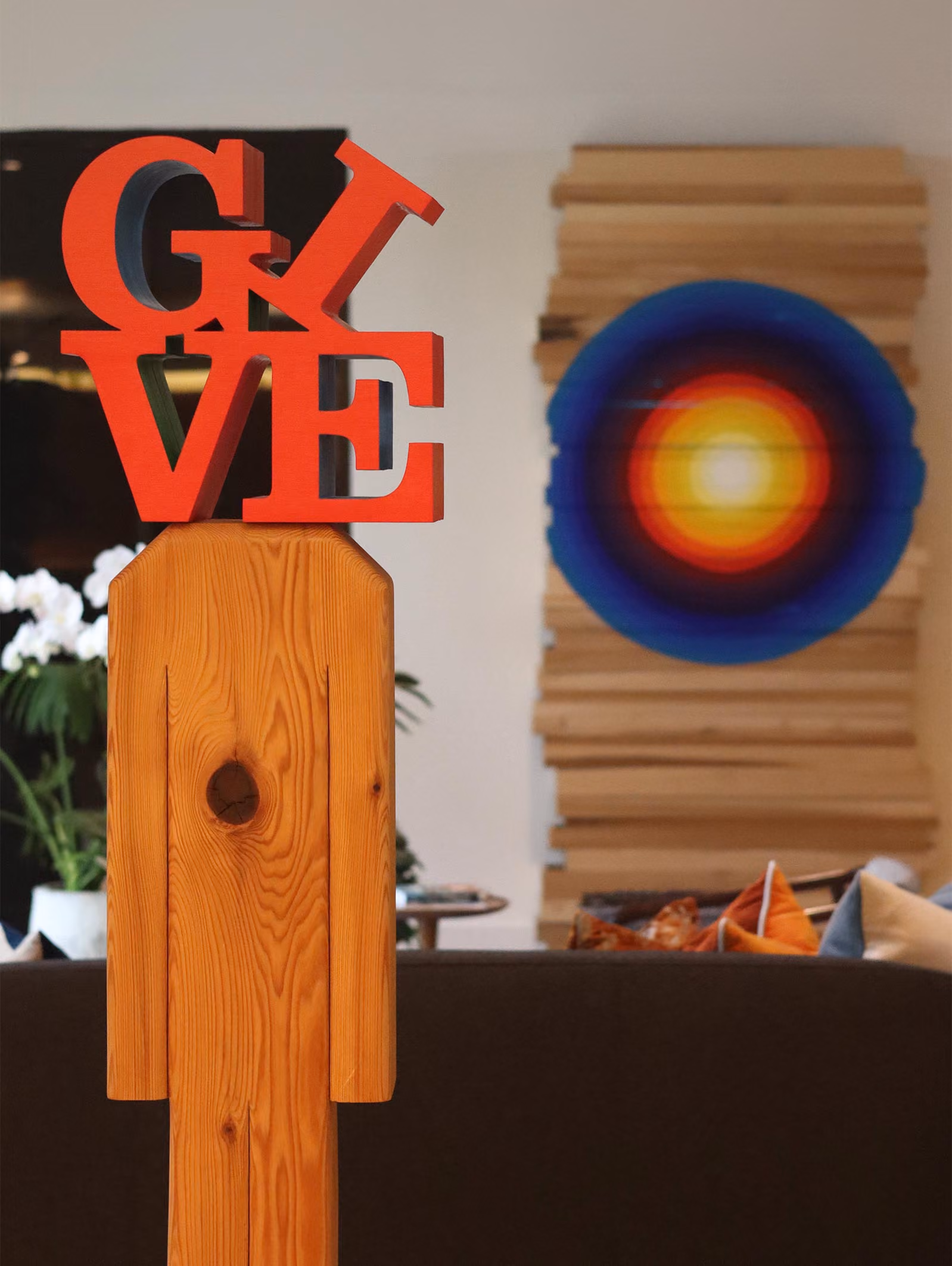 Wooden sculpture resembling a human figure with a hole in the torso, topped with bright red letters spelling GIVE, with the letter I tilted, in a modern indoor setting with colorful cushions and a circular wall art in the background.