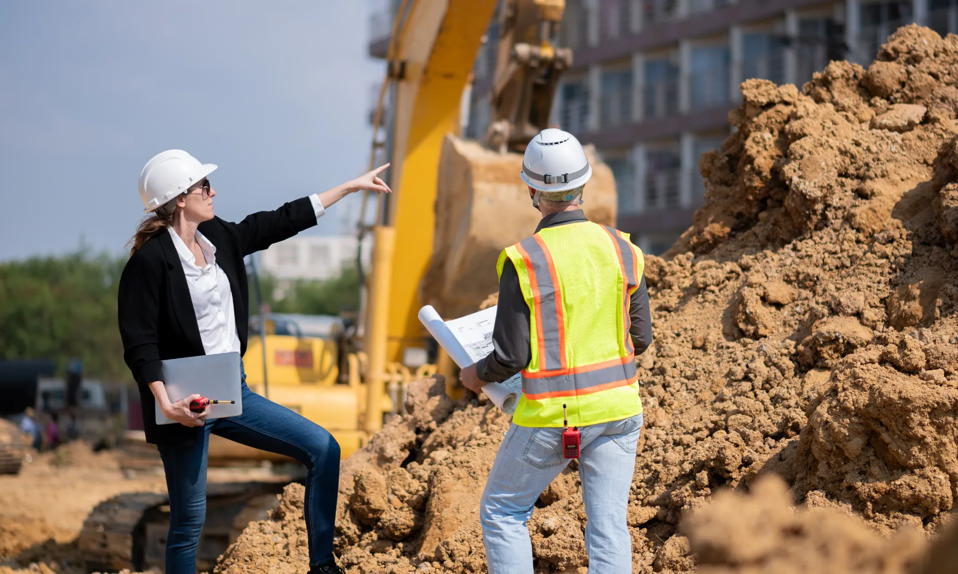 Bauleiterin weist auf Baustelle auf Erdarbeiten, während Ingenieur Pläne prüft – Abstimmung statt Lean-Stillstand.