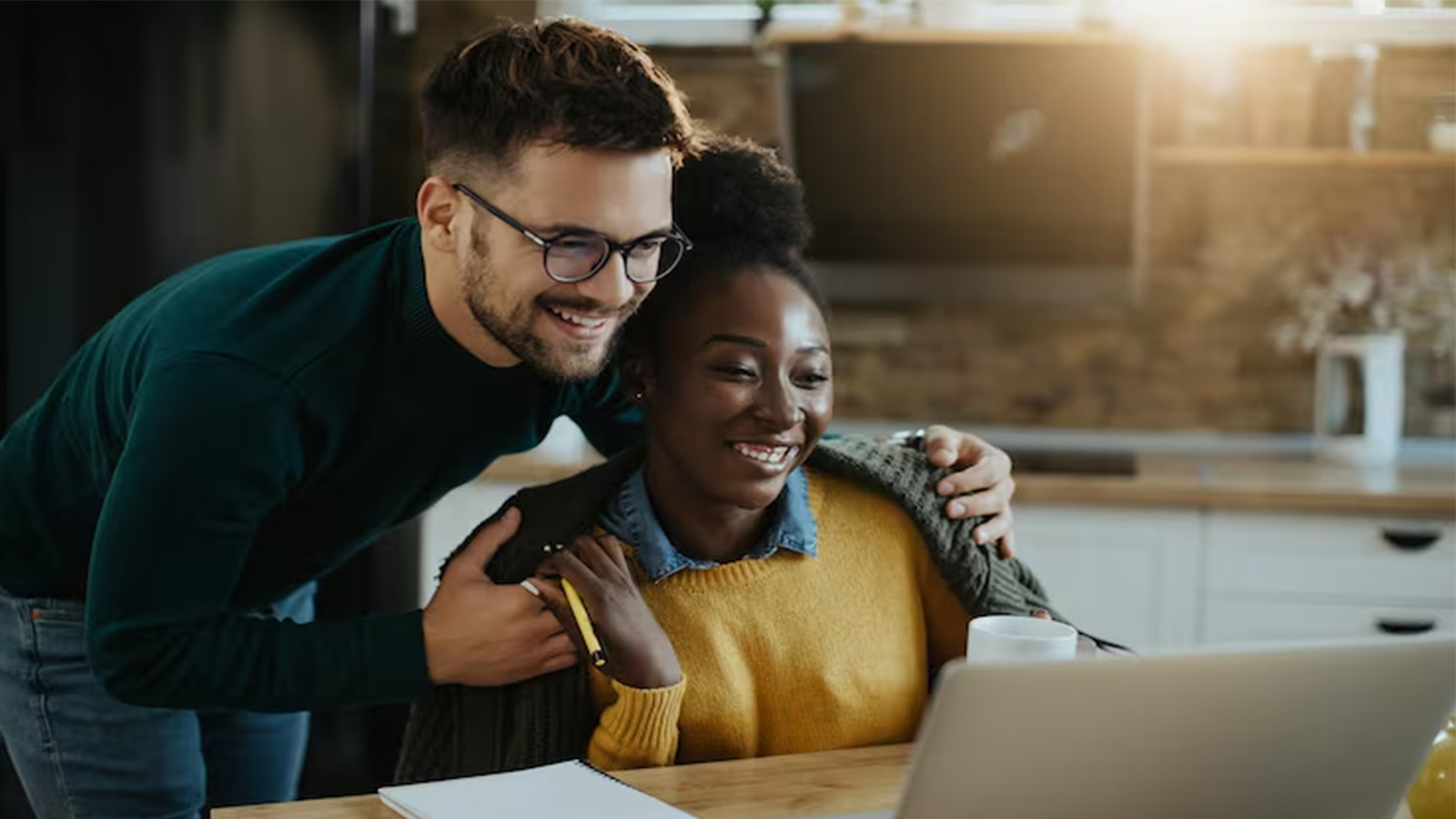 Man embracing woman from behind as they smile while looking at a laptop in a cozy kitchen.