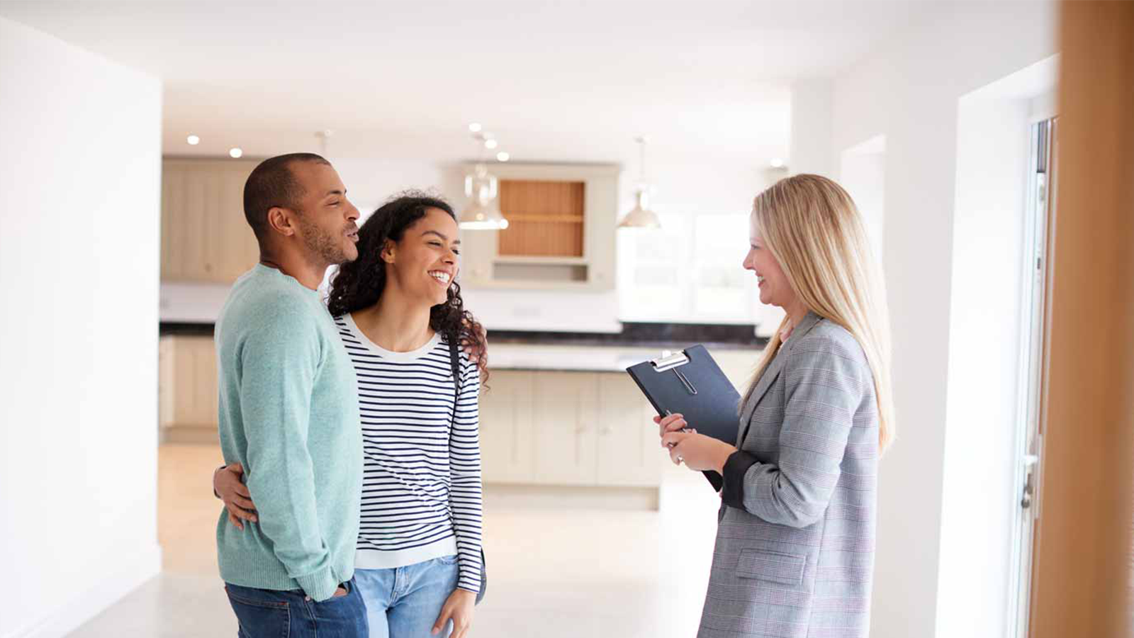 Couple smiling and talking with a woman holding a clipboard in a bright empty kitchen.