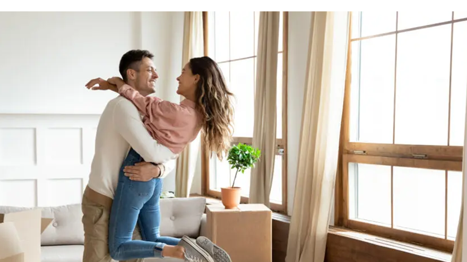 Smiling couple embracing in a bright living room with large windows and a potted plant.