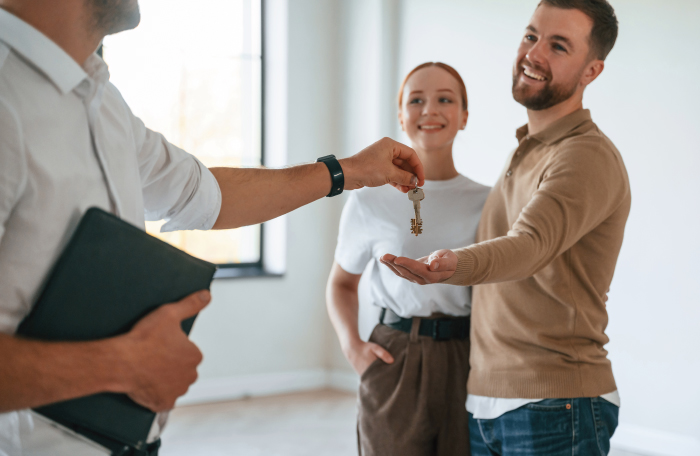 Man handing house key to smiling man while woman stands behind in an empty bright room.