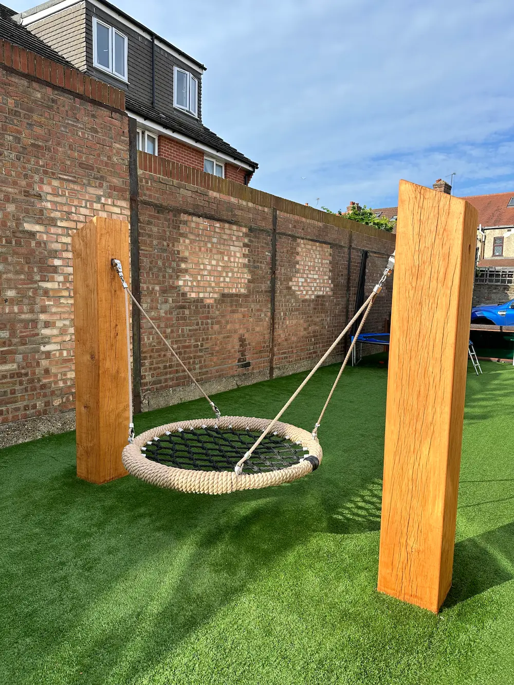 Round basket swing suspended by ropes between two wooden posts over green artificial grass with brick walls and houses in the background.