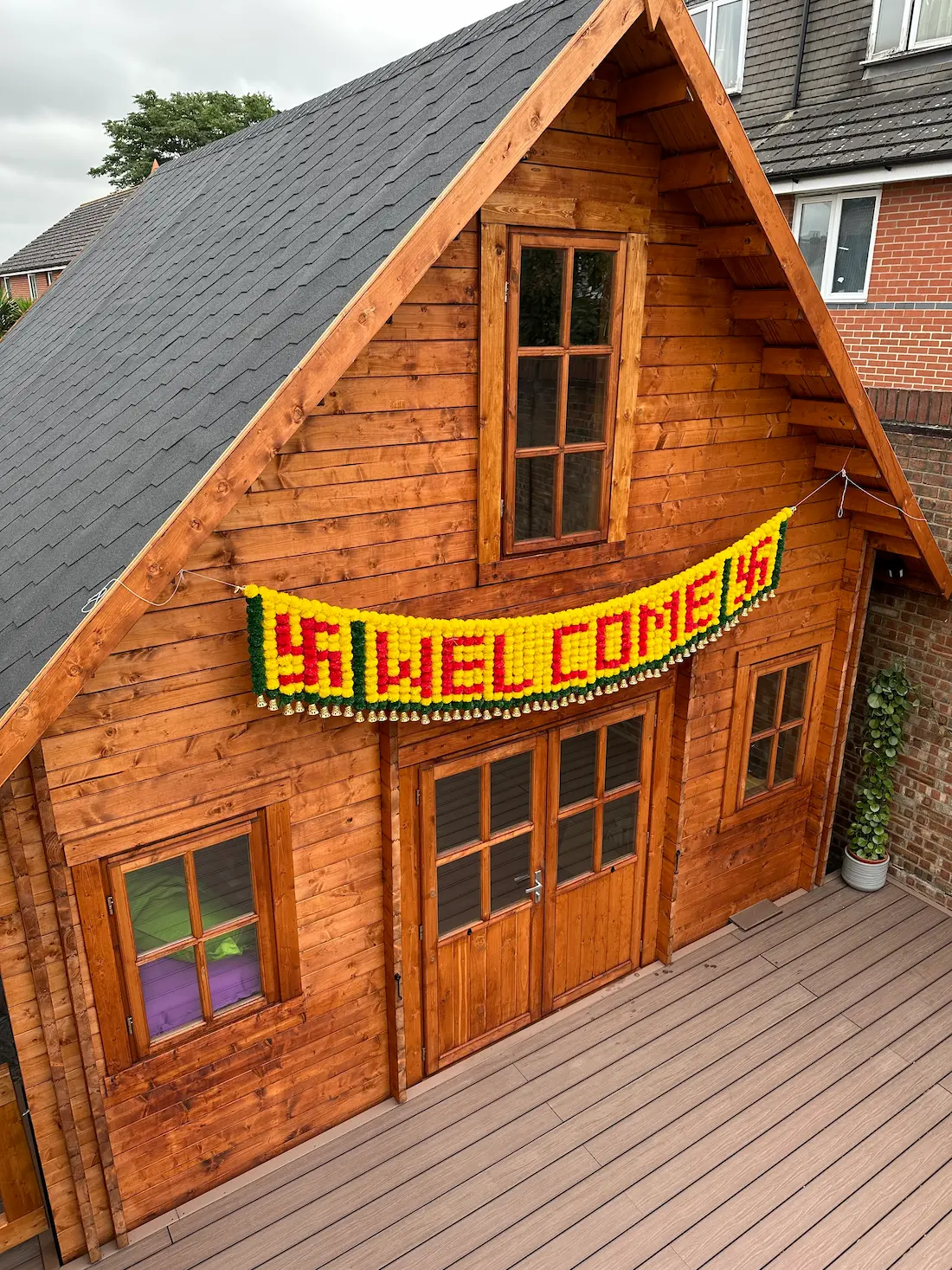 Wooden cabin with a dark shingled roof, featuring a yellow and red welcome banner hanging above the double door on a wooden deck.