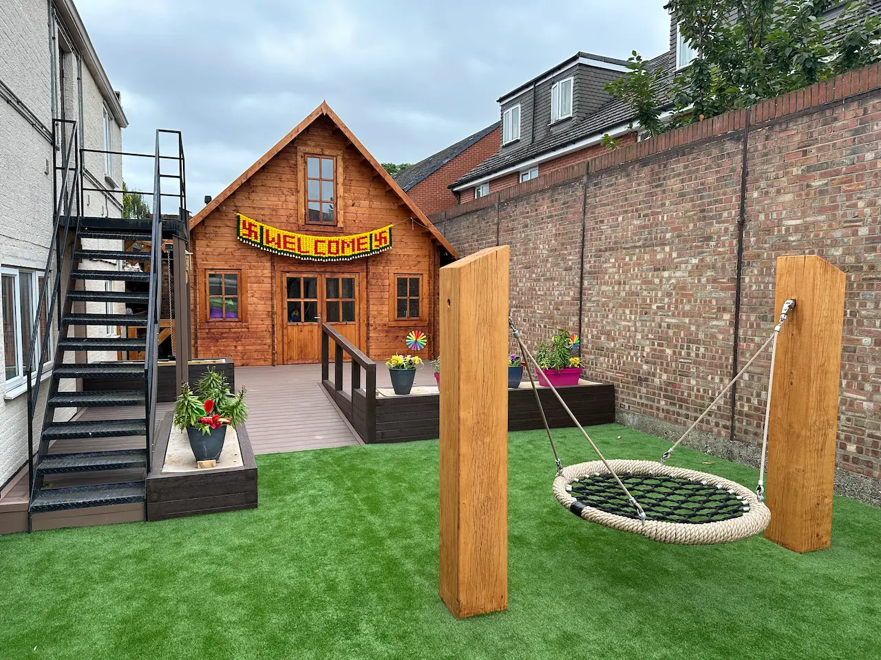 Wooden cabin with a 'WELCOME' sign, a metal staircase, and a circular rope swing on artificial grass in a fenced backyard.