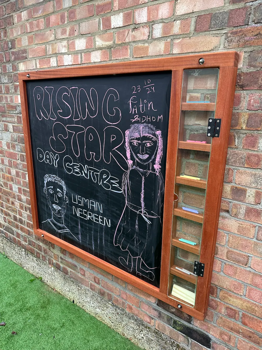 Outdoor chalkboard on brick wall with 'Rising Star Day Centre' written in large letters, a drawing of a girl with braided hair, a man's face sketch, and names Usman Negreen and Fatin Bidhom.
