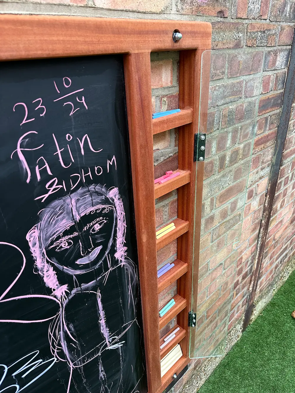 Close-up of a wooden-framed blackboard mounted on a brick wall with pink chalk drawing of a girl and various colored chalk sticks stored in shelves on the frame.