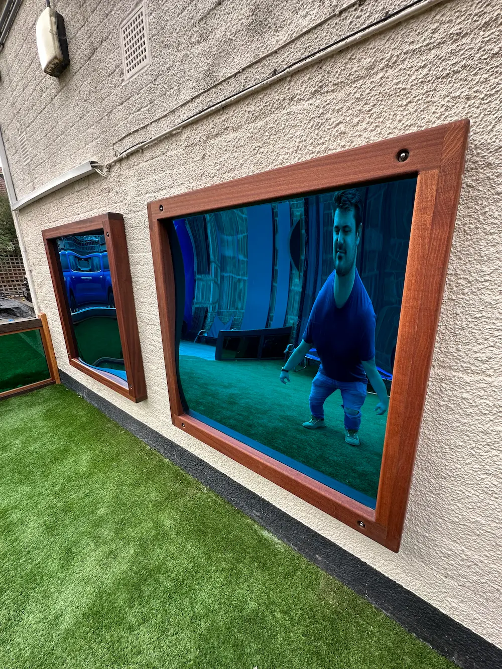 Man distorted in a funhouse mirror on an outdoor wall next to another distorted reflection mirror.