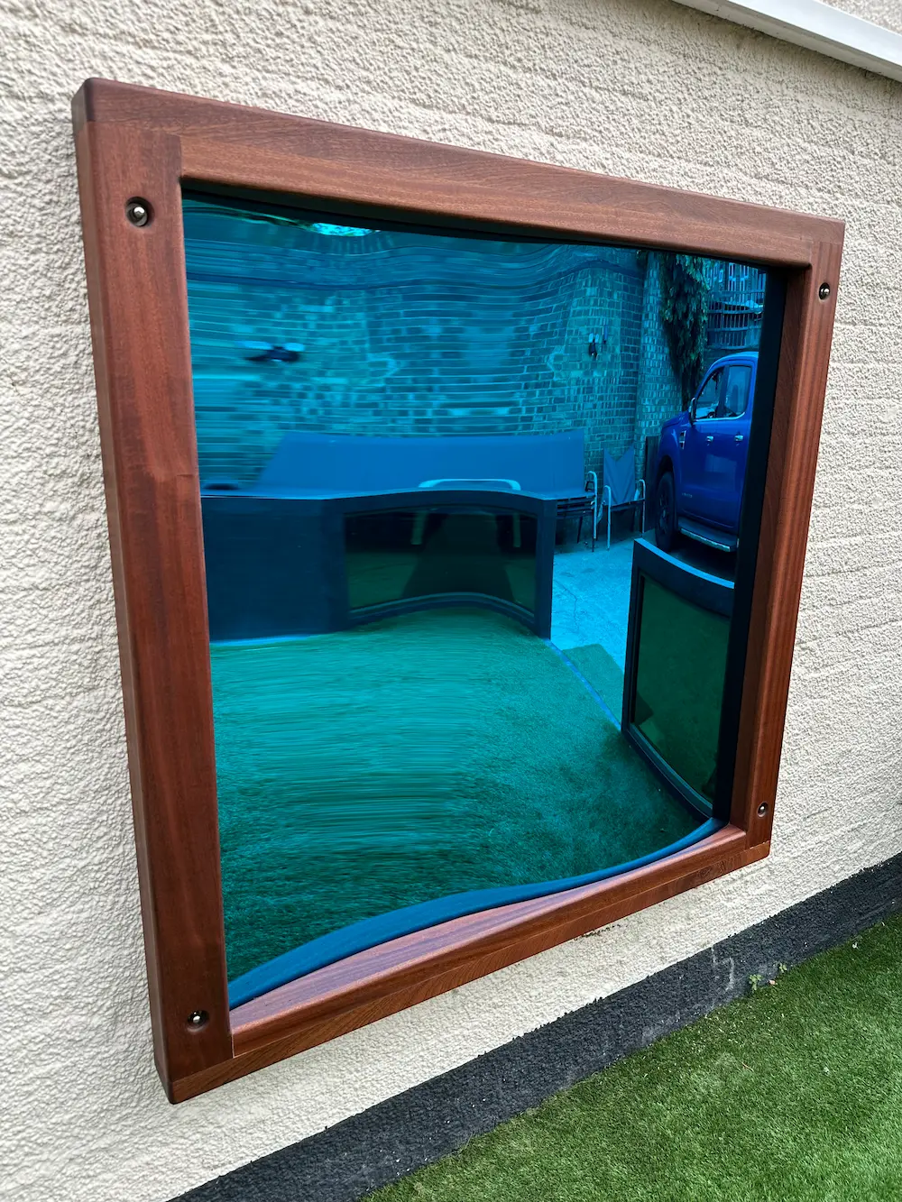 Wall-mounted blue reflective mirror with a wooden frame distorting the reflection of a grassy yard, a brick wall, a blue vehicle, and outdoor furniture.