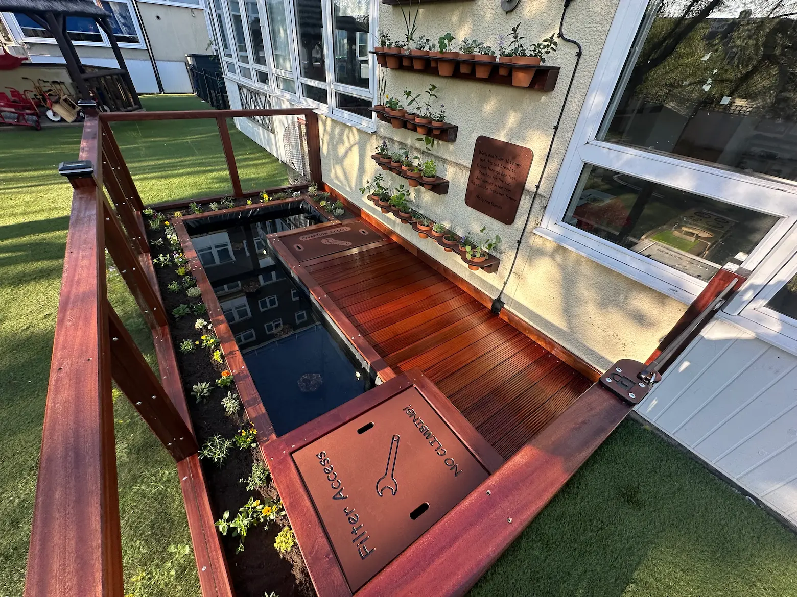 Outdoor eco pond with wooden deck and glass panel railing, surrounded by small plants and wall-mounted potted plants.
