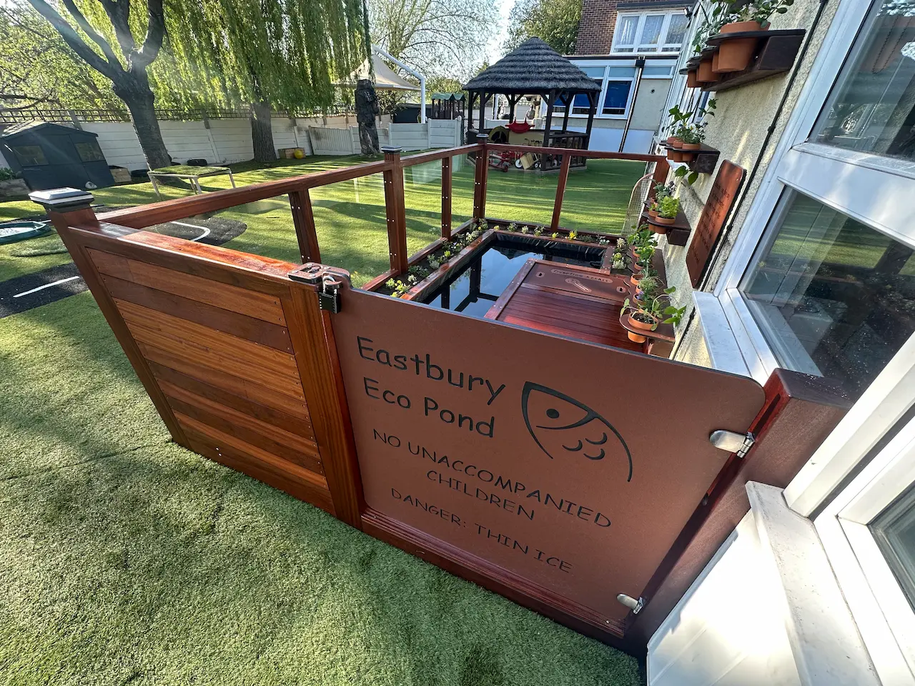 Outdoor eco pond with wooden deck and glass panel railing, surrounded by small plants and wall-mounted potted plants.