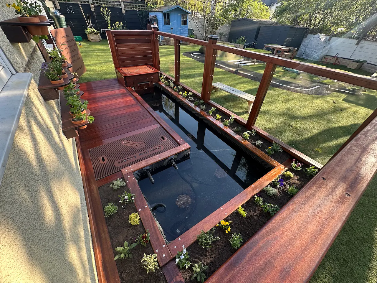 Modern backyard wooden deck with built-in fish pond surrounded by small flowering plants and a glass railing overlooking a garden with lawn and sheds.
