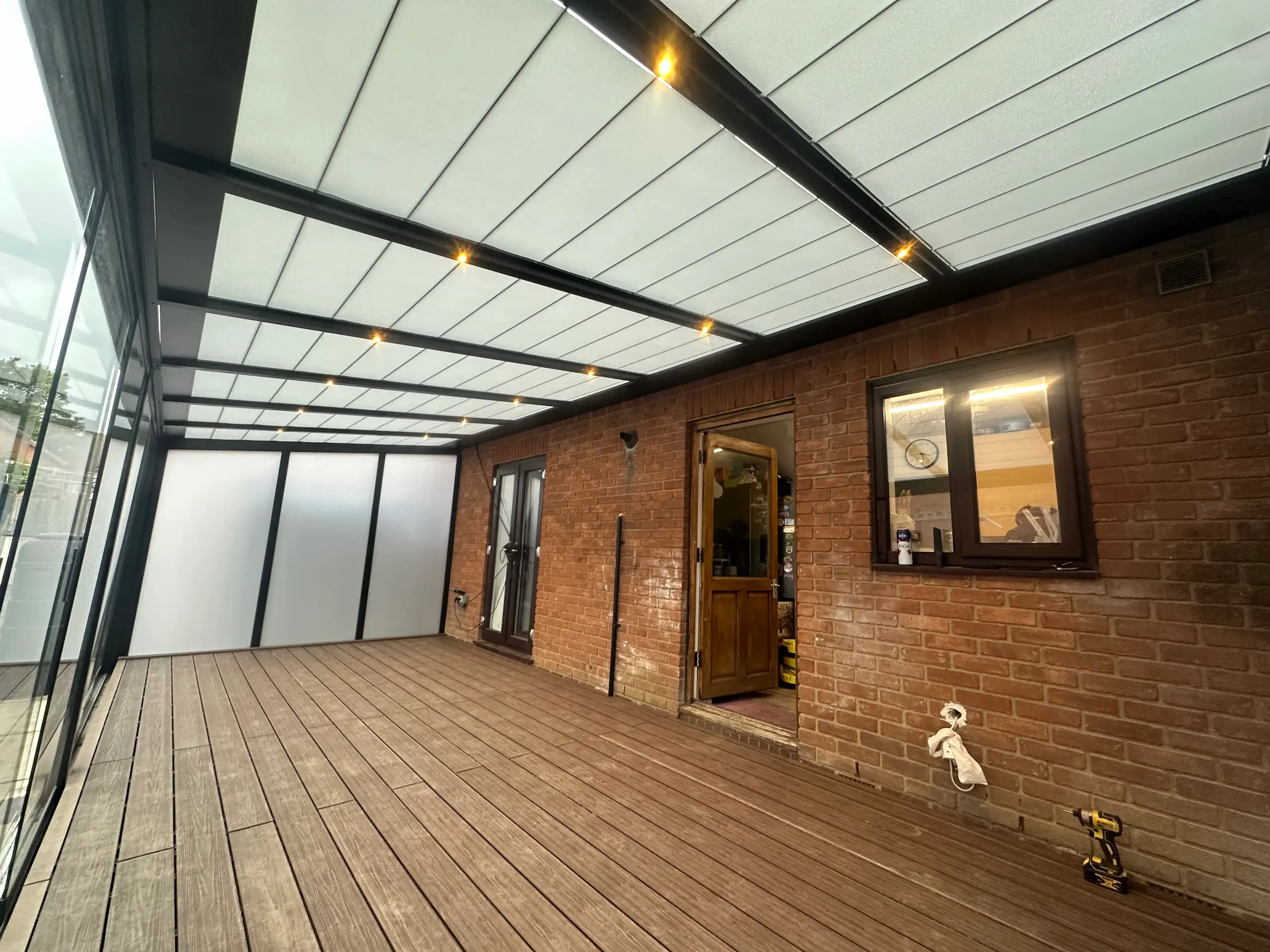 Covered outdoor patio with wood decking, brick wall, frosted glass panels, and ceiling lights.
