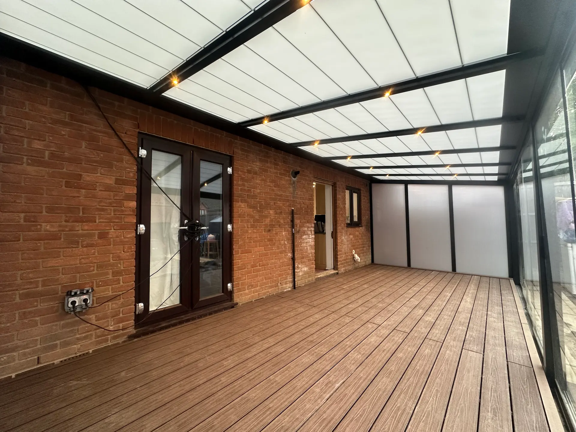 Covered patio with wood-style flooring, brick wall, glass doors, and a translucent roof with recessed lights.
