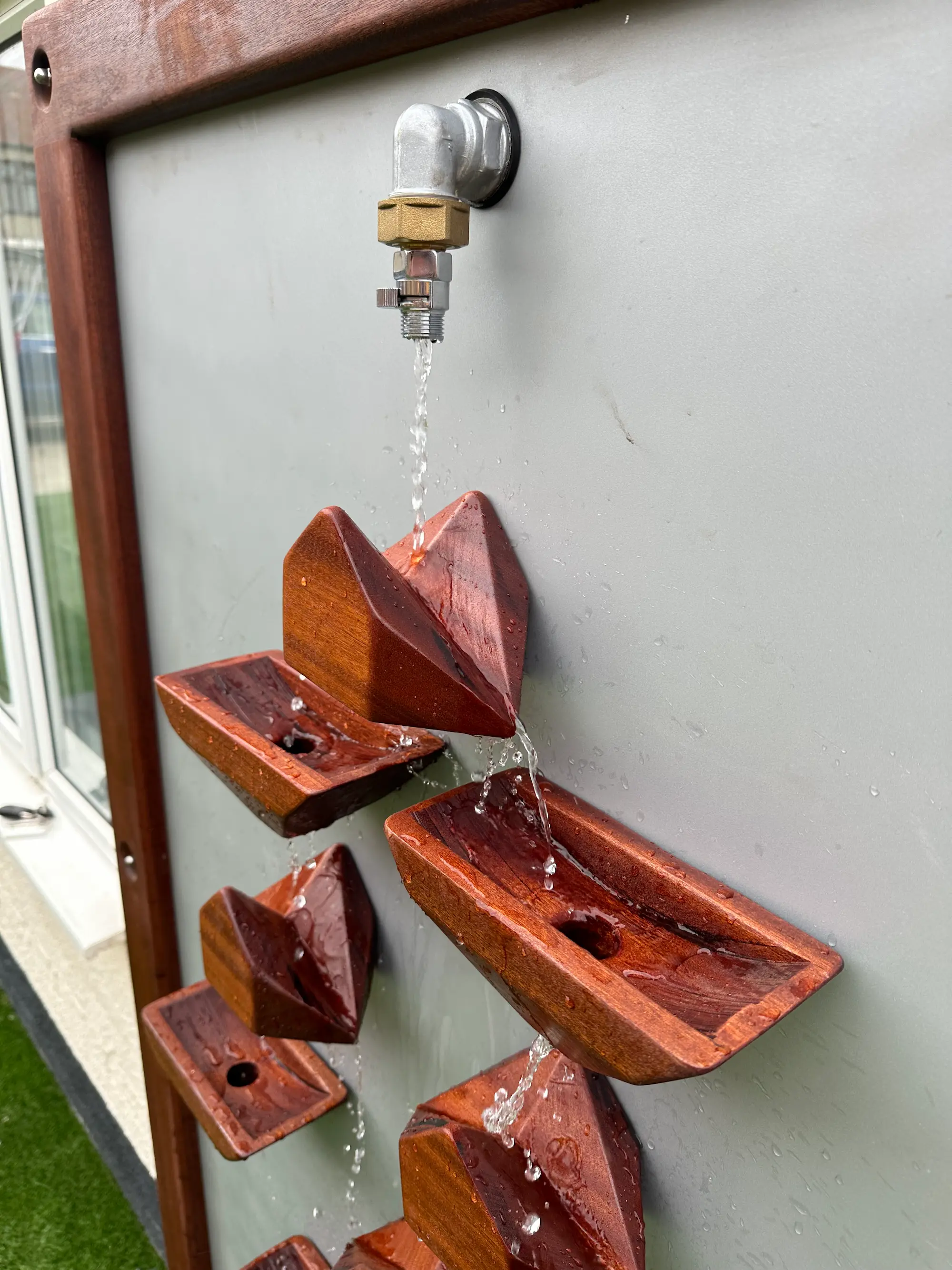 Wall-mounted water feature with a tap pouring water into a series of angled wooden troughs arranged vertically.