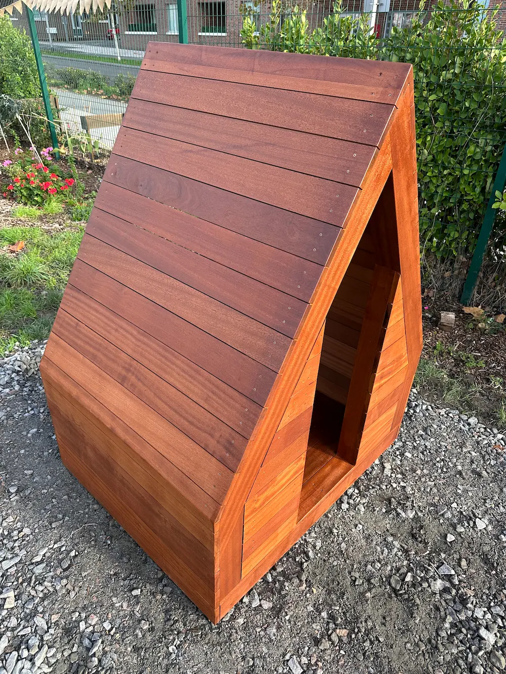 Small wooden A-frame doghouse on gravel with a green fence and plants in the background.