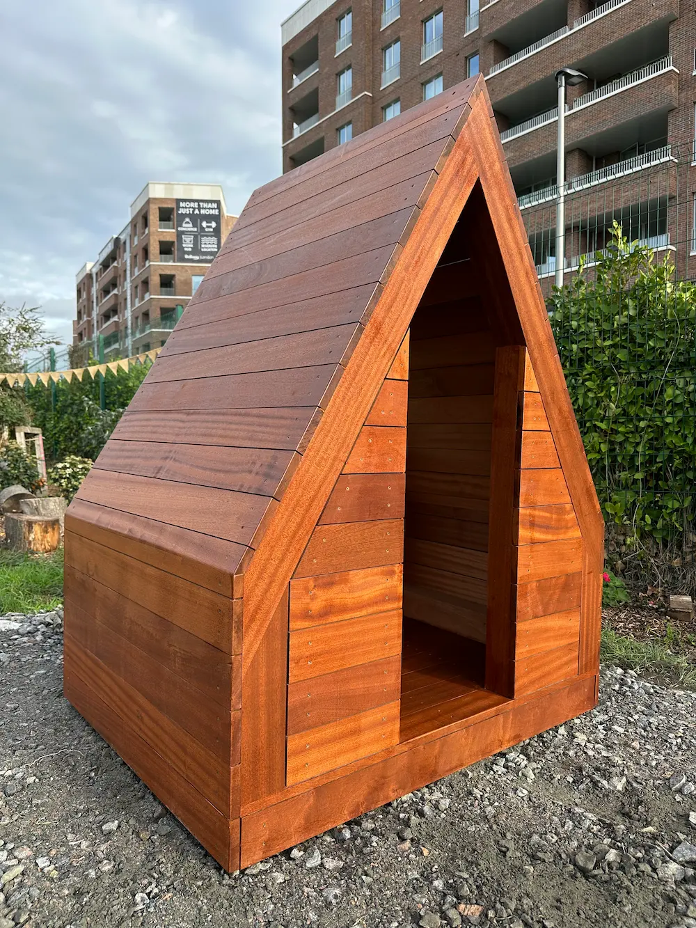 Small wooden A-frame structure resembling a mini cottage or dog house placed outdoors on gravel.
