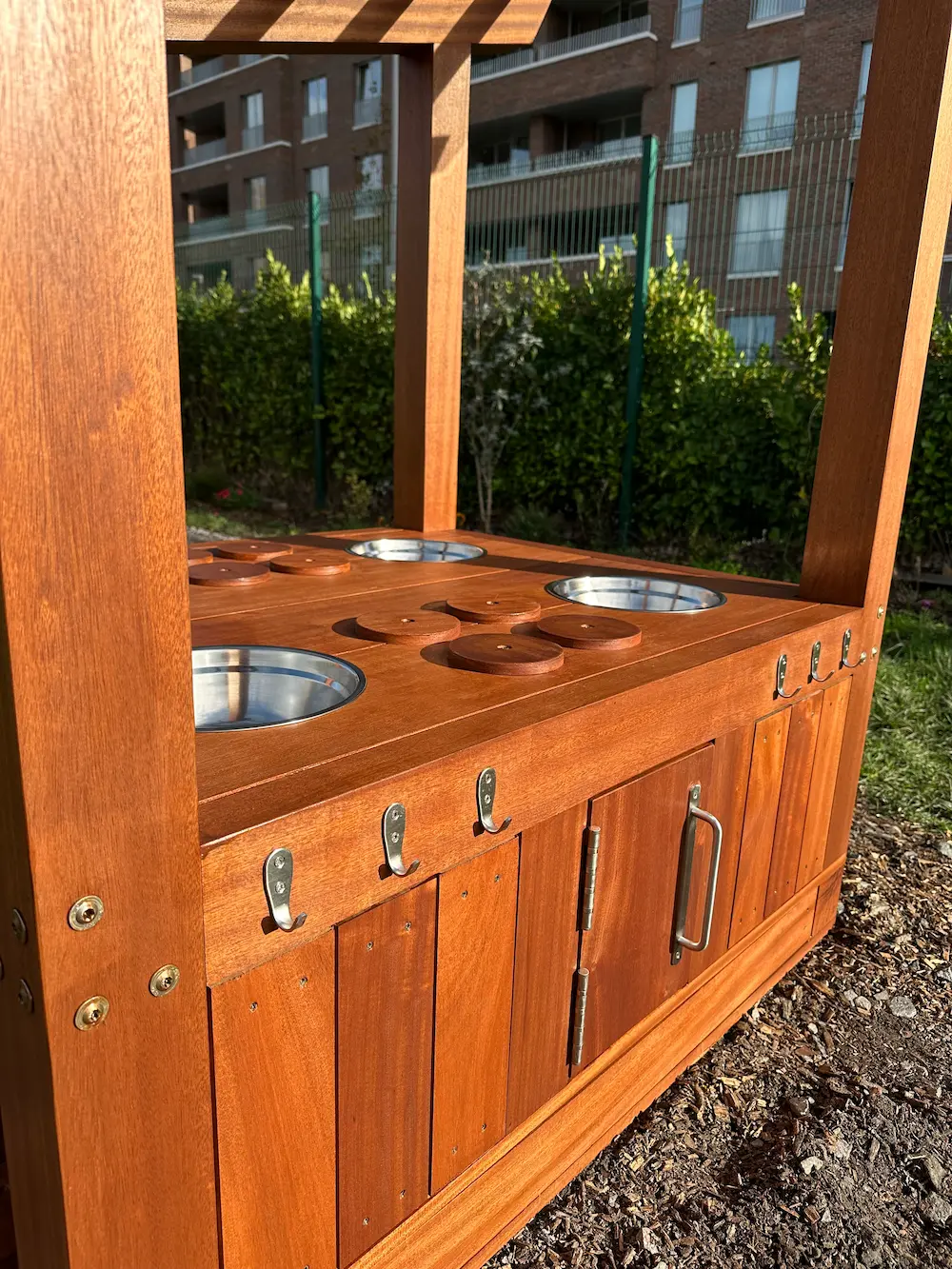 Wooden outdoor play kitchen with metal bowls, wooden lids, and hooks, set in a garden area.