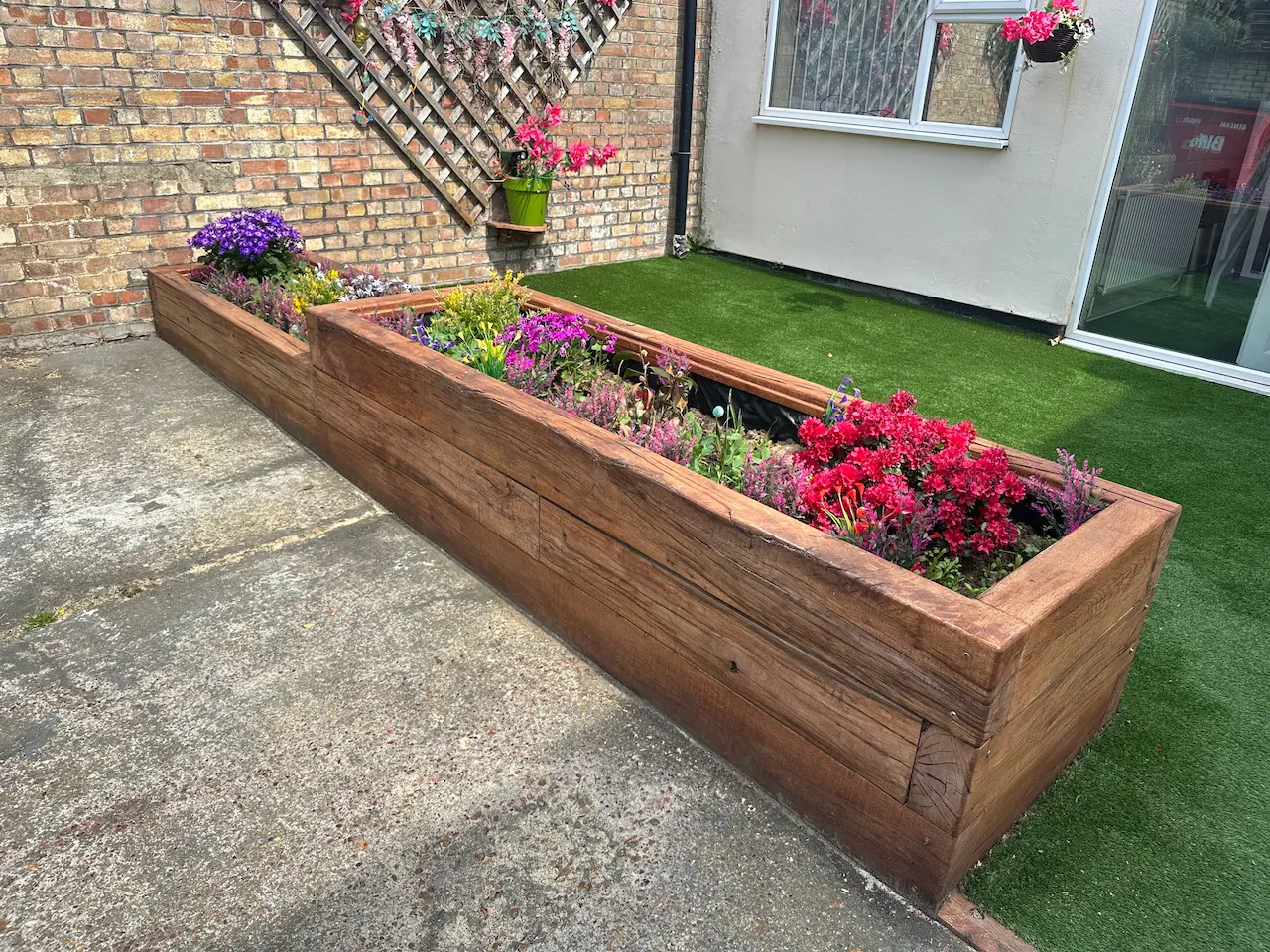 Two large wooden rectangular planters with colorful flowers placed on concrete and artificial grass near a brick wall and white house exterior.