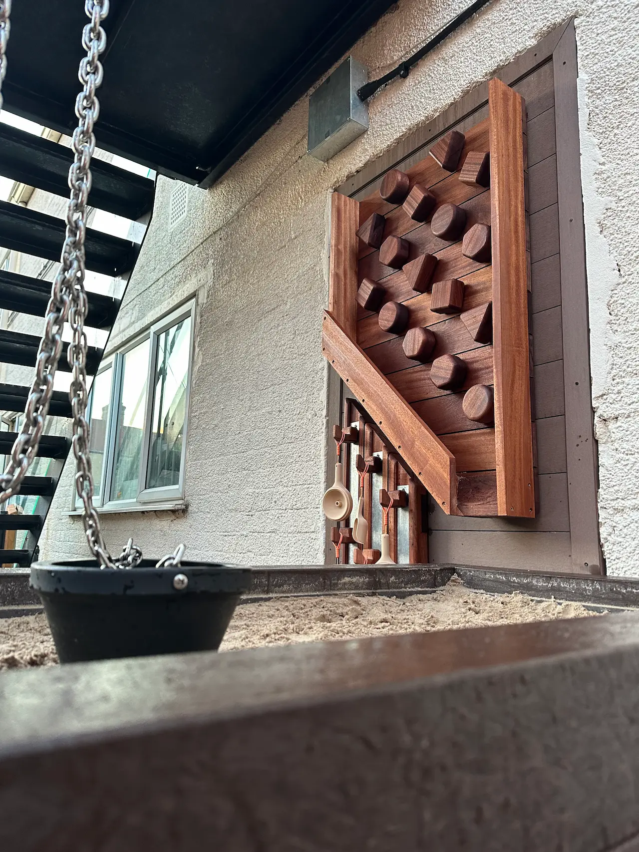 Close-up of a black bucket swing hanging by chains above a sandbox, with a wooden climbing wall featuring various shapes mounted on a stucco wall in the background.