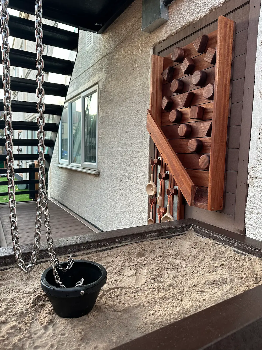 A sandbox with a black bucket swing hanging from metal chains and a wooden tactile panel with various geometric shapes mounted on a wall next to a door.