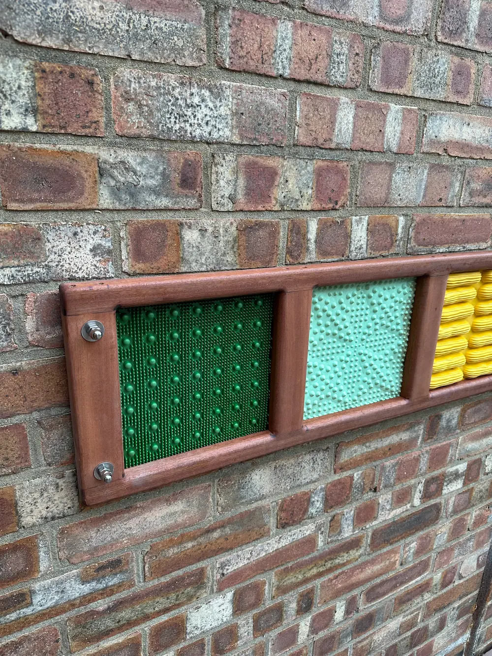 Close-up of a tactile sensory panel mounted on a brick wall with green, light blue, and yellow textured sections framed in wood.