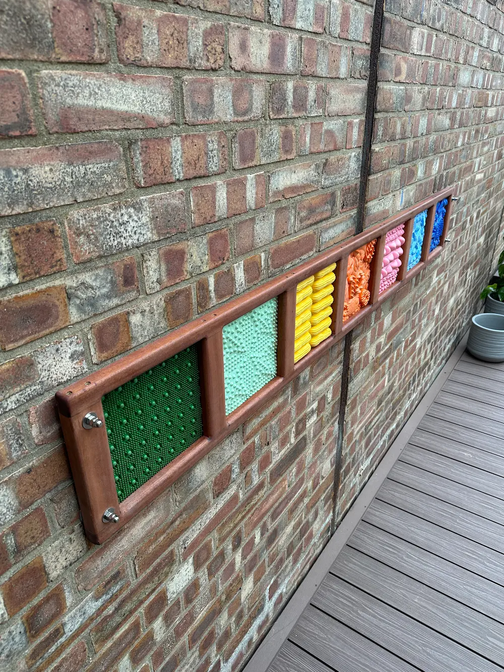 Wall-mounted sensory panel with seven colorful textured squares in green, light blue, yellow, orange, pink, and two shades of blue.
