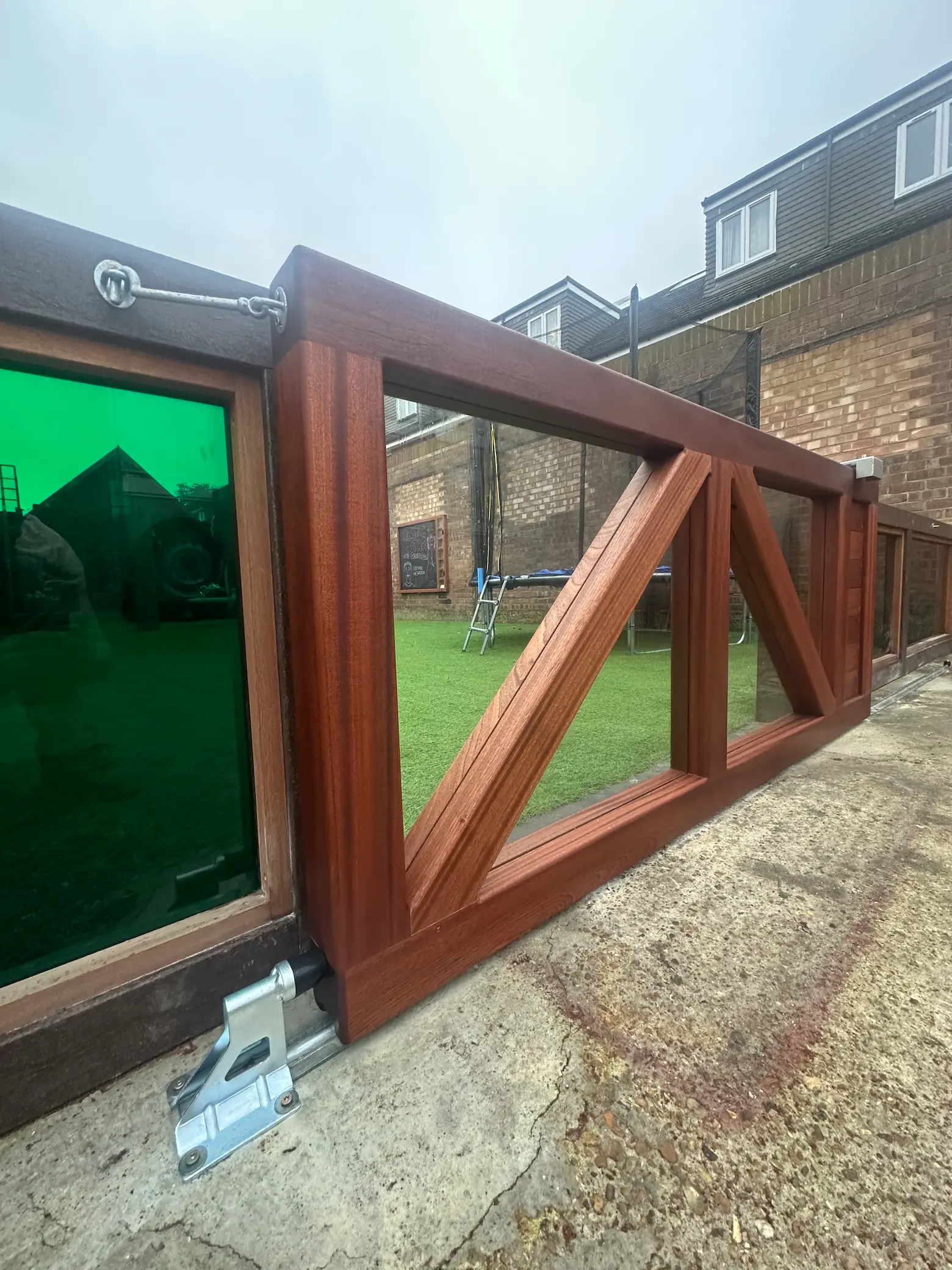 Sliding wooden gate with diagonal support beams set on a concrete driveway next to a green-tinted glass panel.