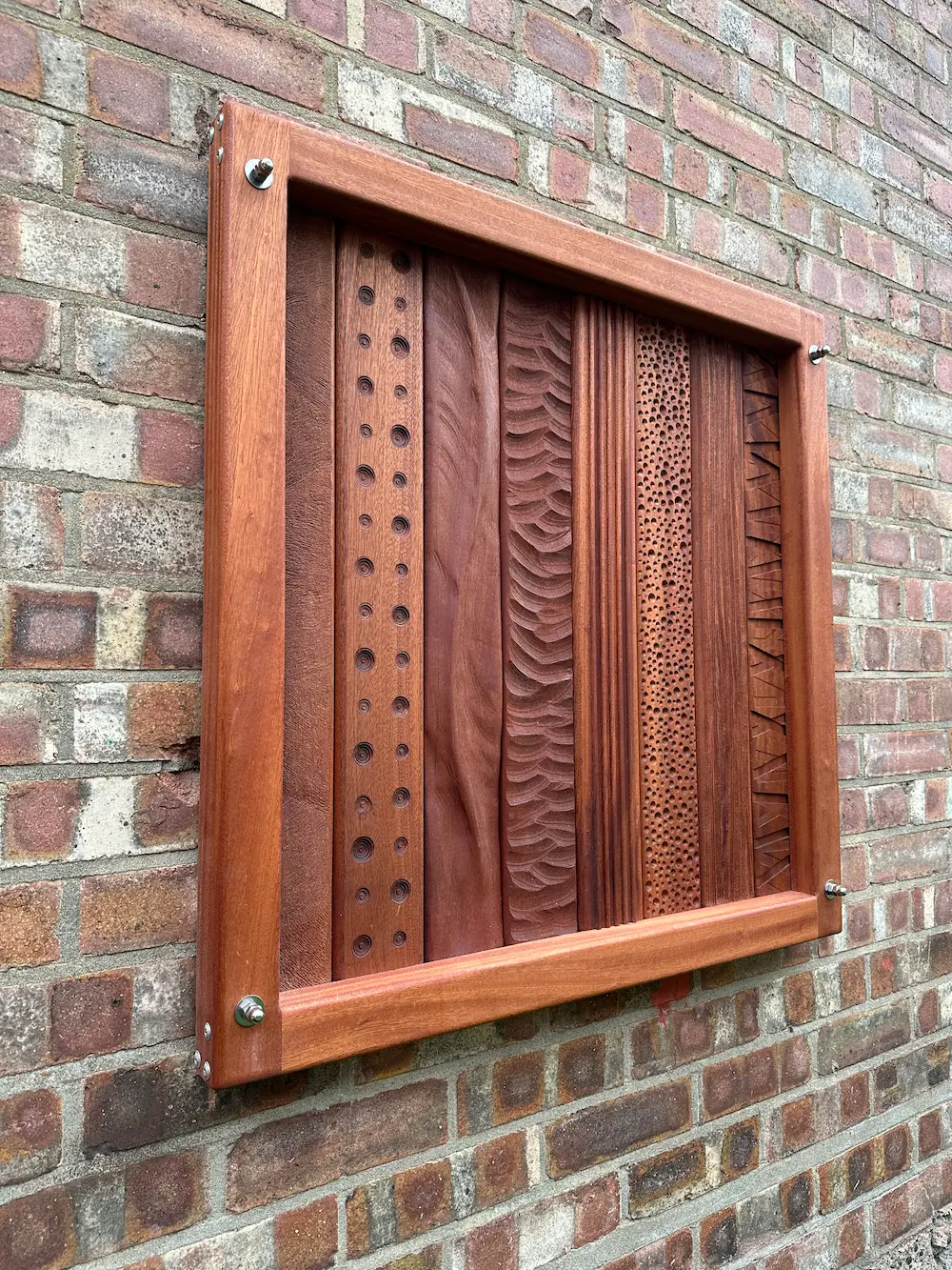 Wooden tactile panel with five different textured vertical strips mounted on an exterior brick wall.
