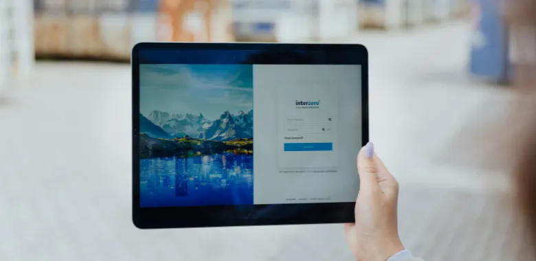 A photograph of a person holding a tablet. The tablet displays a login screen for "interzero" with fields for username and password against a background image of a serene mountain landscape reflected in a lake. The person's hand has a manicured nail, suggesting a detail-oriented and professional individual. The setting appears to be outdoors, with blurred street scenes in the background indicating an urban environment.