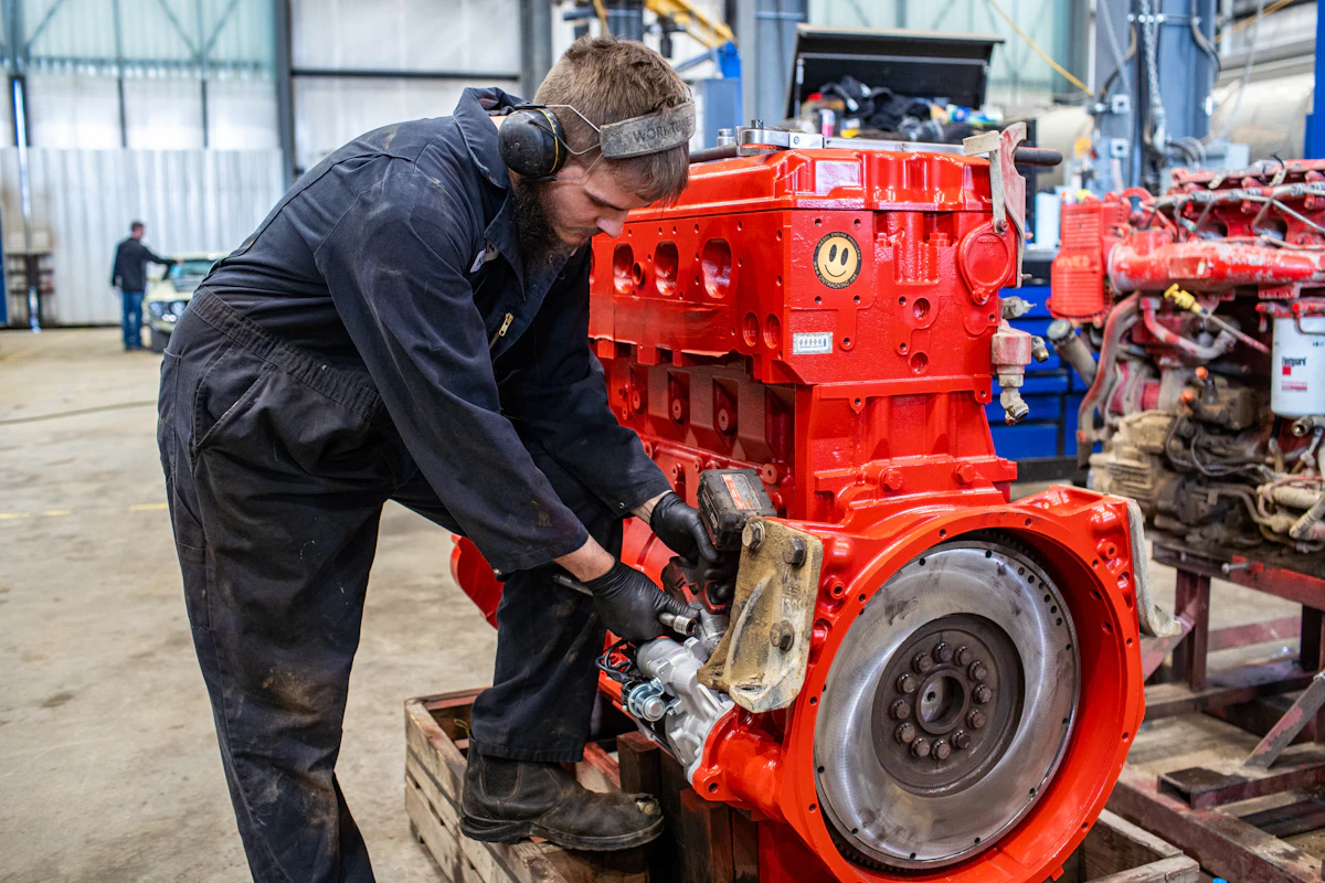 Mechanic wearing coveralls, gloves, and hearing protection working on a large red diesel engine inside a truck repair shop.