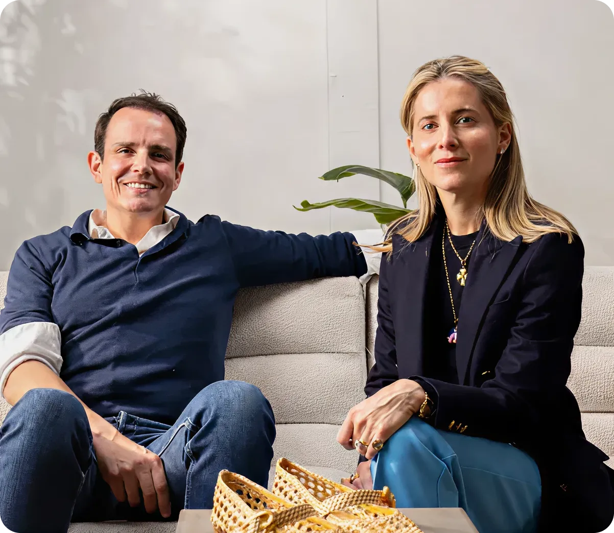 Smiling man in a dark blue shirt with rolled-up sleeves and woman with blonde hair wearing a black blazer and blue pants sitting on a beige sofa with woven baskets on the table in front.