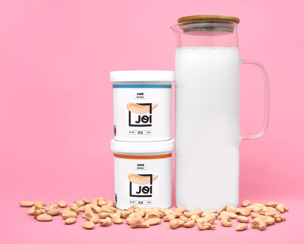 Smiling man in a dark blue shirt with rolled-up sleeves and woman with blonde hair wearing a black blazer and blue pants sitting on a beige sofa with woven baskets on the table in front.