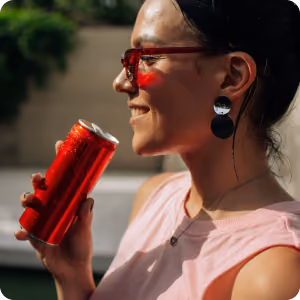Smiling woman wearing red sunglasses holding a red beverage can near her mouth.