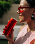 Smiling woman wearing red sunglasses holding a red beverage can near her mouth.