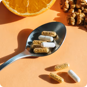 Spoon containing several white and yellow capsules with scattered capsules and orange slice in the background.