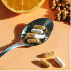Spoon containing several white and yellow capsules with scattered capsules and orange slice in the background.