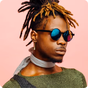 Man with dreadlocks and dyed tips wearing reflective sunglasses, a green sweater, and a light scarf against a pink background.