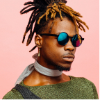 Man with dreadlocks and dyed tips wearing reflective sunglasses, a green sweater, and a light scarf against a pink background.