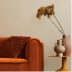 Orange velvet sofa with a dark brown cushion next to a side table holding two brown vases with dried flowers.