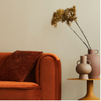 Orange velvet sofa with a dark brown cushion next to a side table holding two brown vases with dried flowers.