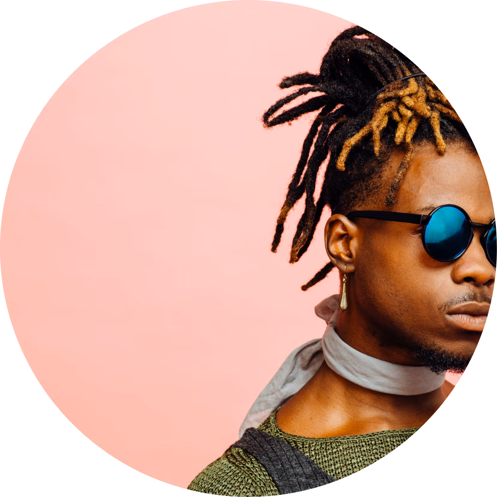 Portrait of a man with dreadlocks wearing round blue sunglasses, a green knit top, gray scarf, and dangling earring against a pink background.