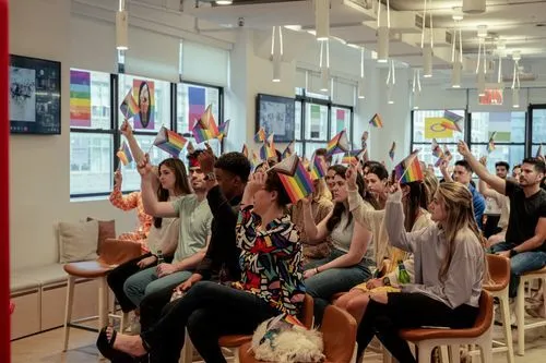 A diverse group of people seated indoors raising small pride flags, with pride-themed posters on the windows behind them.